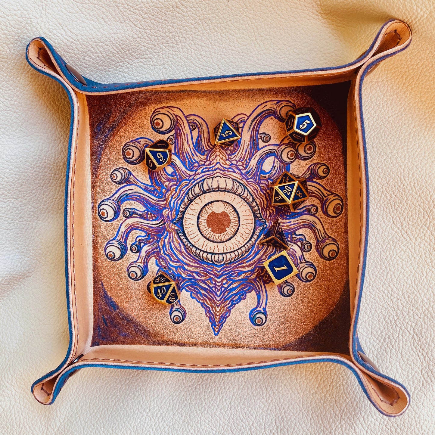 SteamVoyage - vassoio lancia dadi in cuoio dipinto a mano rivestito con pelle blu, dotato di quattro bottoni angolari per la chiusura e cucitura sul bordo. Il vassoio lancia dadi è decorato con l'immagine di un beholder, mostro tipico di Dungeons and dragons, dipinto con colori vivaci: viola e blu