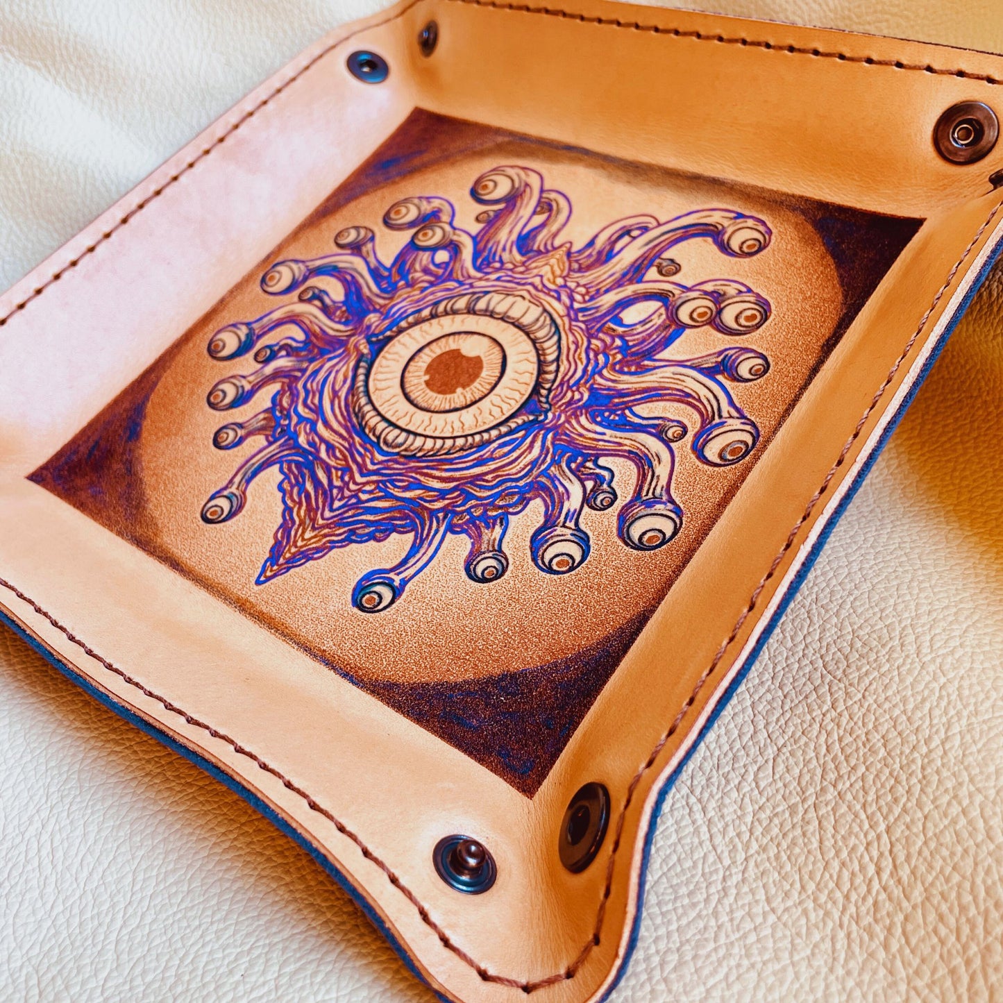 SteamVoyage - vassoio lancia dadi in cuoio dipinto a mano rivestito con pelle blu, dotato di quattro bottoni angolari per la chiusura e cucitura sul bordo. Il vassoio lancia dadi è decorato con l'immagine di un beholder, mostro tipico di Dungeons and dragons, dipinto con colori vivaci: viola e blu