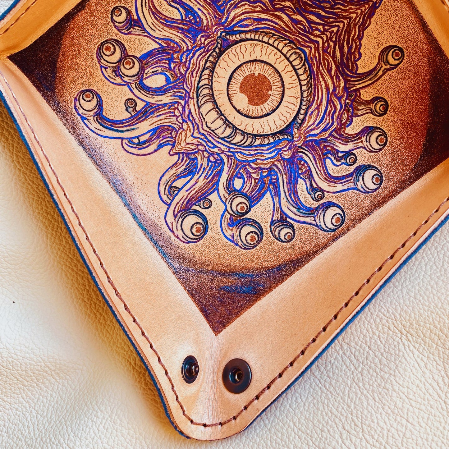 SteamVoyage - vassoio lancia dadi in cuoio dipinto a mano rivestito con pelle blu, dotato di quattro bottoni angolari per la chiusura e cucitura sul bordo. Il vassoio lancia dadi è decorato con l'immagine di un beholder, mostro tipico di Dungeons and dragons, dipinto con colori vivaci: viola e blu