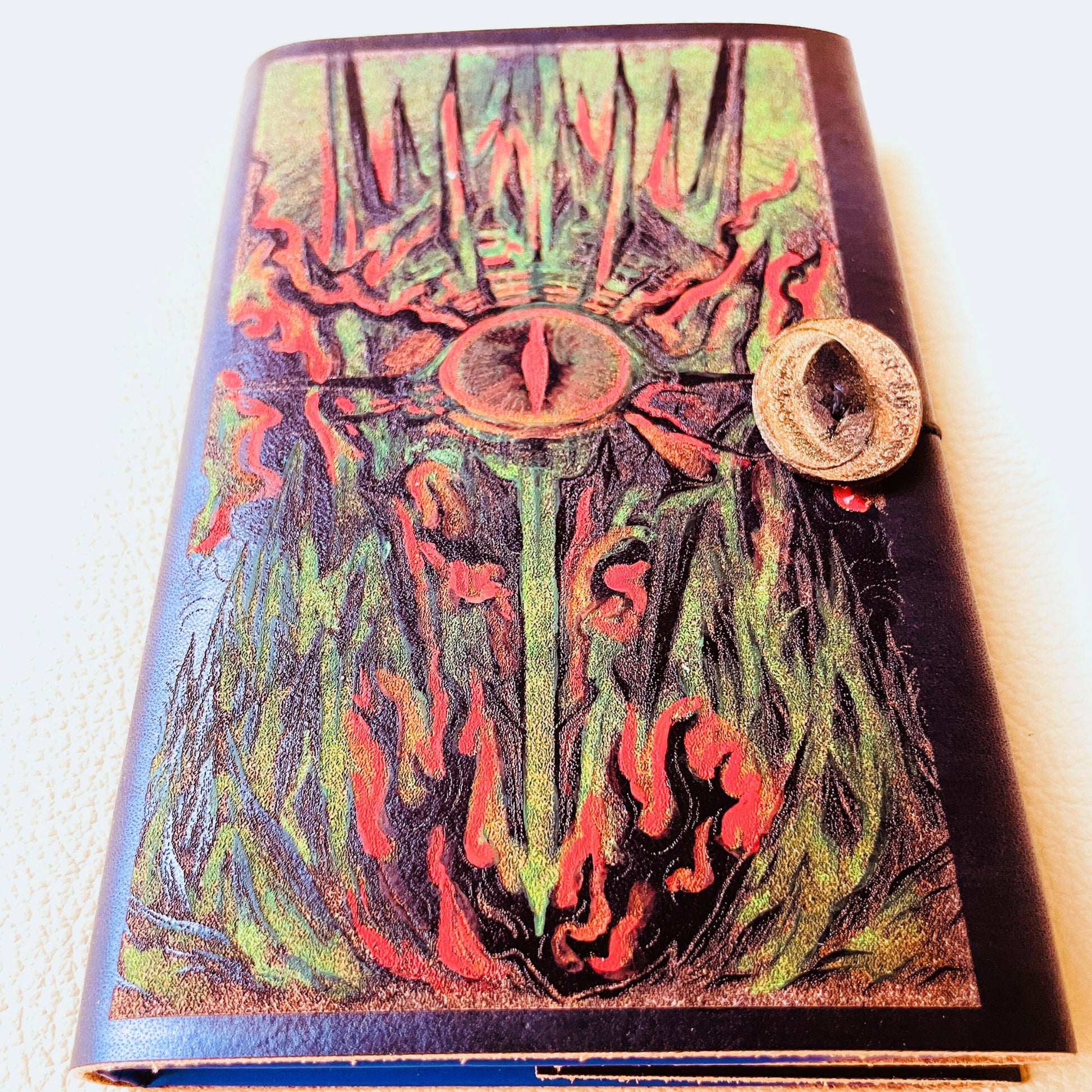 SteamVoyage - copertina in cuoio tinto di nero per agenda A6 con incisione a laser della Torre di Sauron dipinta a mano con colori fluo e cordino per la chiusura ornato da due anelli del potere in cuoio dorato