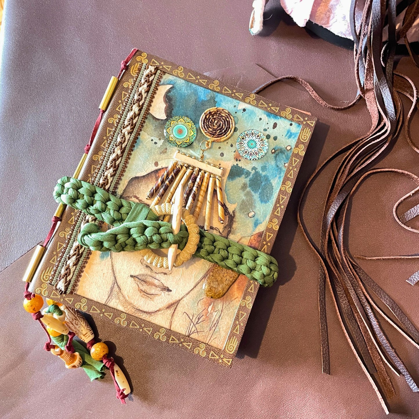 SteamVoyage - libro magico della sciamana in legno decorato con la tecnica del decoupage, rivestito in pelle marrone incisa al laser con fantasie africane dorate, con applicazioni in stoffa e perline e fettuccia per la chiusura in lycra lavorata all'uncinetto. All'interno, stampati su carta di pregio, si trovano preghiere e invocazioni, ricette per elisir magici e indicazioni per rituali arcani, oltre ad annotazioni personali della maga.