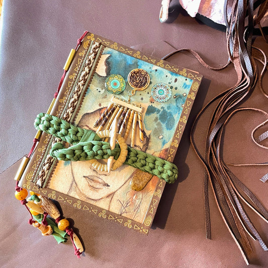 SteamVoyage - libro magico della sciamana in legno decorato con la tecnica del decoupage, rivestito in pelle marrone incisa al laser con fantasie africane dorate, con applicazioni in stoffa e perline e fettuccia per la chiusura in lycra lavorata all'uncinetto. All'interno, stampati su carta di pregio, si trovano preghiere e invocazioni, ricette per elisir magici e indicazioni per rituali arcani, oltre ad annotazioni personali della maga.