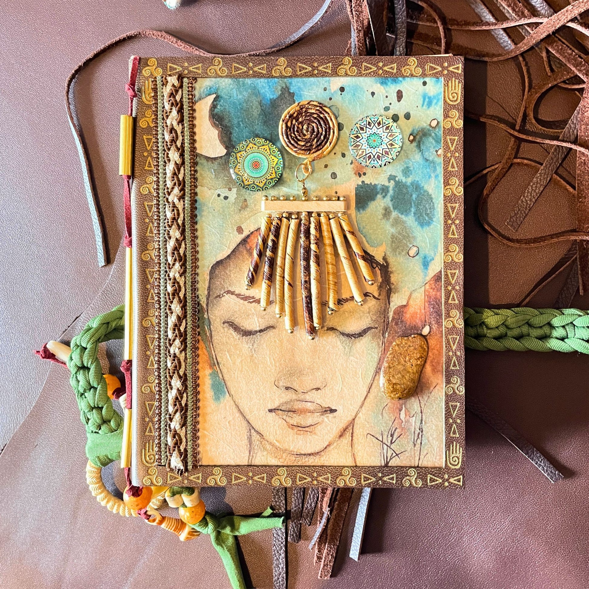 SteamVoyage - libro magico della sciamana in legno decorato con la tecnica del decoupage, rivestito in pelle marrone incisa al laser con fantasie africane dorate, con applicazioni in stoffa e perline e fettuccia per la chiusura in lycra lavorata all'uncinetto. All'interno, stampati su carta di pregio, si trovano preghiere e invocazioni, ricette per elisir magici e indicazioni per rituali arcani, oltre ad annotazioni personali della maga.