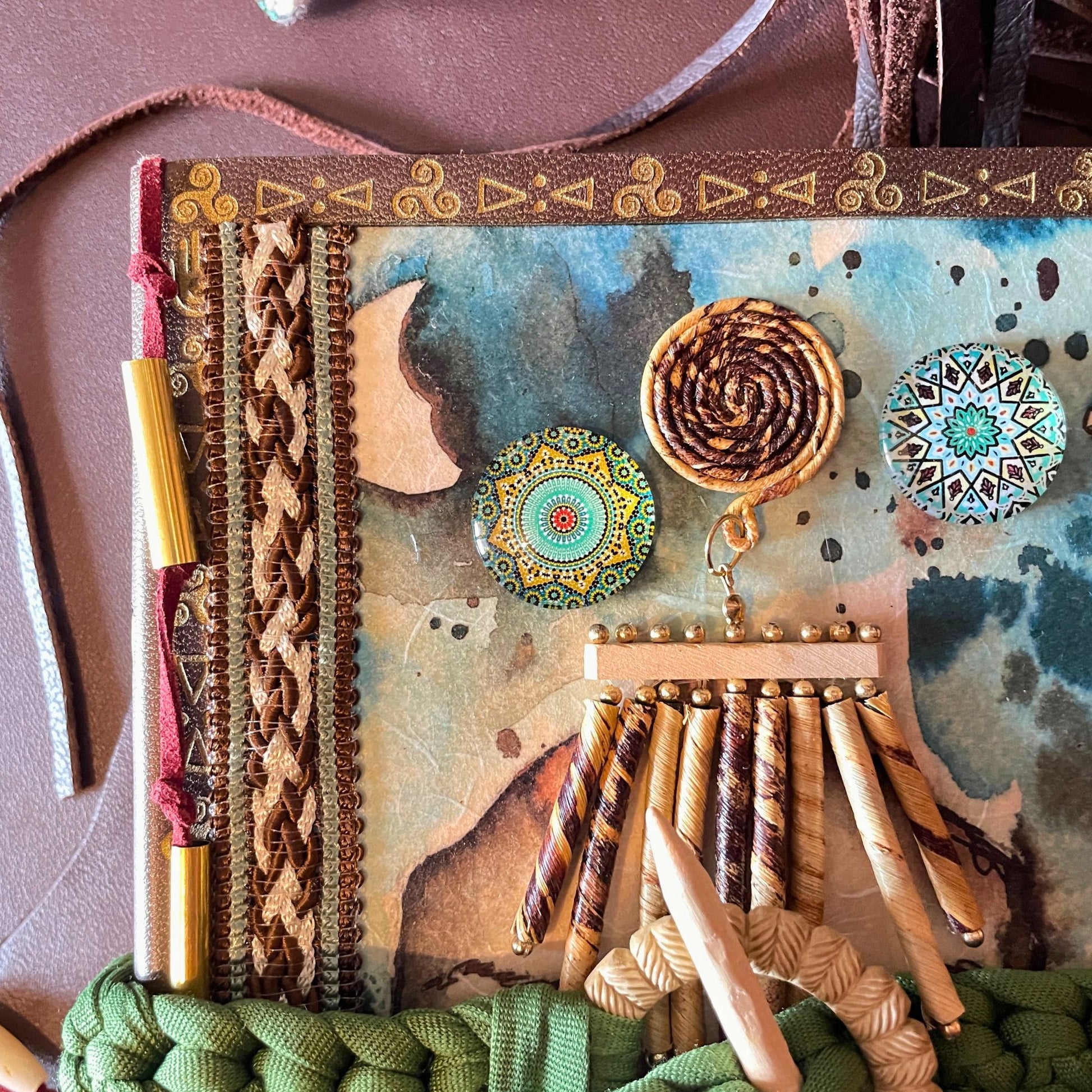 SteamVoyage - libro magico della sciamana in legno decorato con la tecnica del decoupage, rivestito in pelle marrone incisa al laser con fantasie africane dorate, con applicazioni in stoffa e perline e fettuccia per la chiusura in lycra lavorata all'uncinetto. All'interno, stampati su carta di pregio, si trovano preghiere e invocazioni, ricette per elisir magici e indicazioni per rituali arcani, oltre ad annotazioni personali della maga.