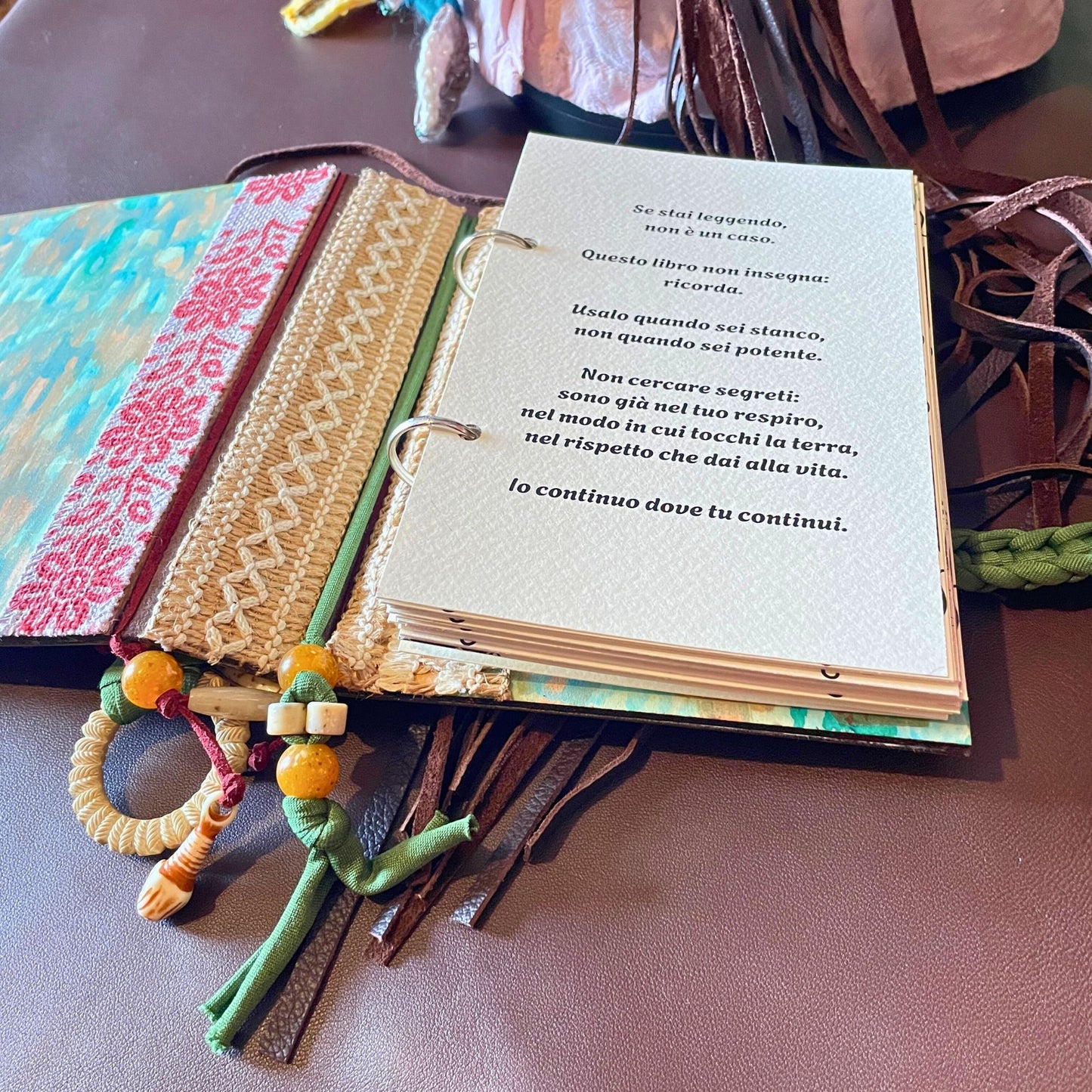 SteamVoyage - libro magico della sciamana in legno decorato con la tecnica del decoupage, rivestito in pelle marrone incisa al laser con fantasie africane dorate, con applicazioni in stoffa e perline e fettuccia per la chiusura in lycra lavorata all'uncinetto. All'interno, stampati su carta di pregio, si trovano preghiere e invocazioni, ricette per elisir magici e indicazioni per rituali arcani, oltre ad annotazioni personali della maga.