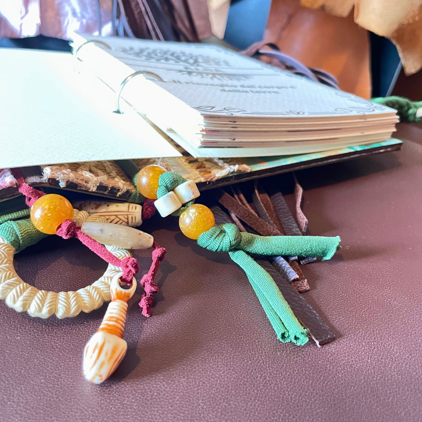 SteamVoyage - libro magico della sciamana in legno decorato con la tecnica del decoupage, rivestito in pelle marrone incisa al laser con fantasie africane dorate, con applicazioni in stoffa e perline e fettuccia per la chiusura in lycra lavorata all'uncinetto. All'interno, stampati su carta di pregio, si trovano preghiere e invocazioni, ricette per elisir magici e indicazioni per rituali arcani, oltre ad annotazioni personali della maga.