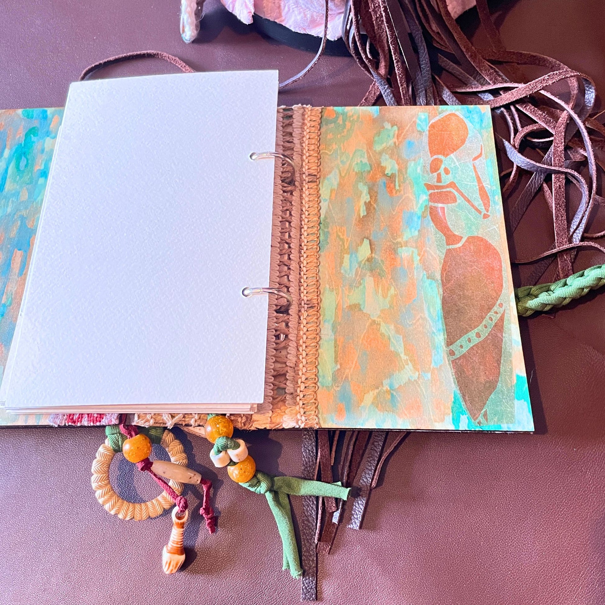 SteamVoyage - libro magico della sciamana in legno decorato con la tecnica del decoupage, rivestito in pelle marrone incisa al laser con fantasie africane dorate, con applicazioni in stoffa e perline e fettuccia per la chiusura in lycra lavorata all'uncinetto. All'interno, stampati su carta di pregio, si trovano preghiere e invocazioni, ricette per elisir magici e indicazioni per rituali arcani, oltre ad annotazioni personali della maga.