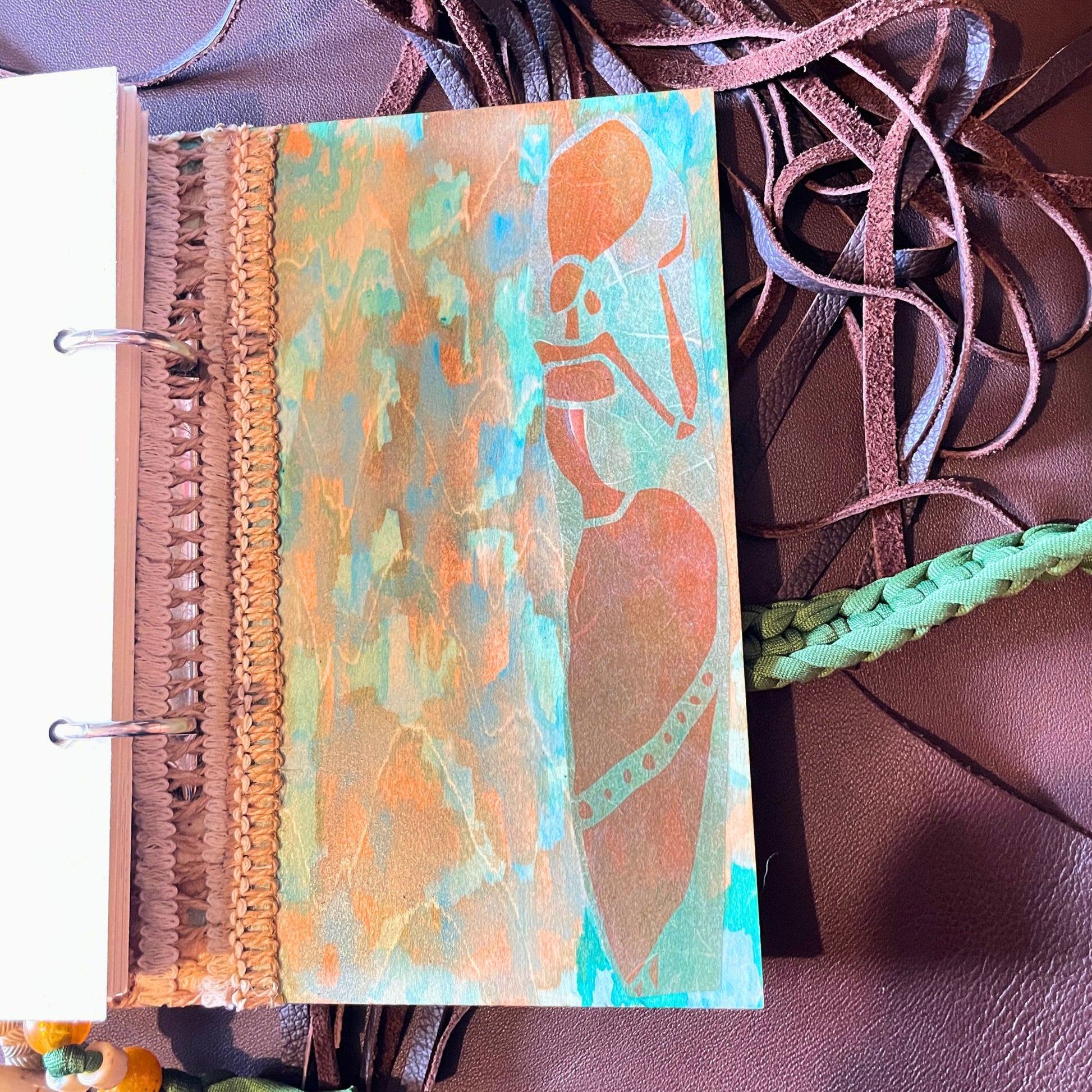 SteamVoyage - libro magico della sciamana in legno decorato con la tecnica del decoupage, rivestito in pelle marrone incisa al laser con fantasie africane dorate, con applicazioni in stoffa e perline e fettuccia per la chiusura in lycra lavorata all'uncinetto. All'interno, stampati su carta di pregio, si trovano preghiere e invocazioni, ricette per elisir magici e indicazioni per rituali arcani, oltre ad annotazioni personali della maga.