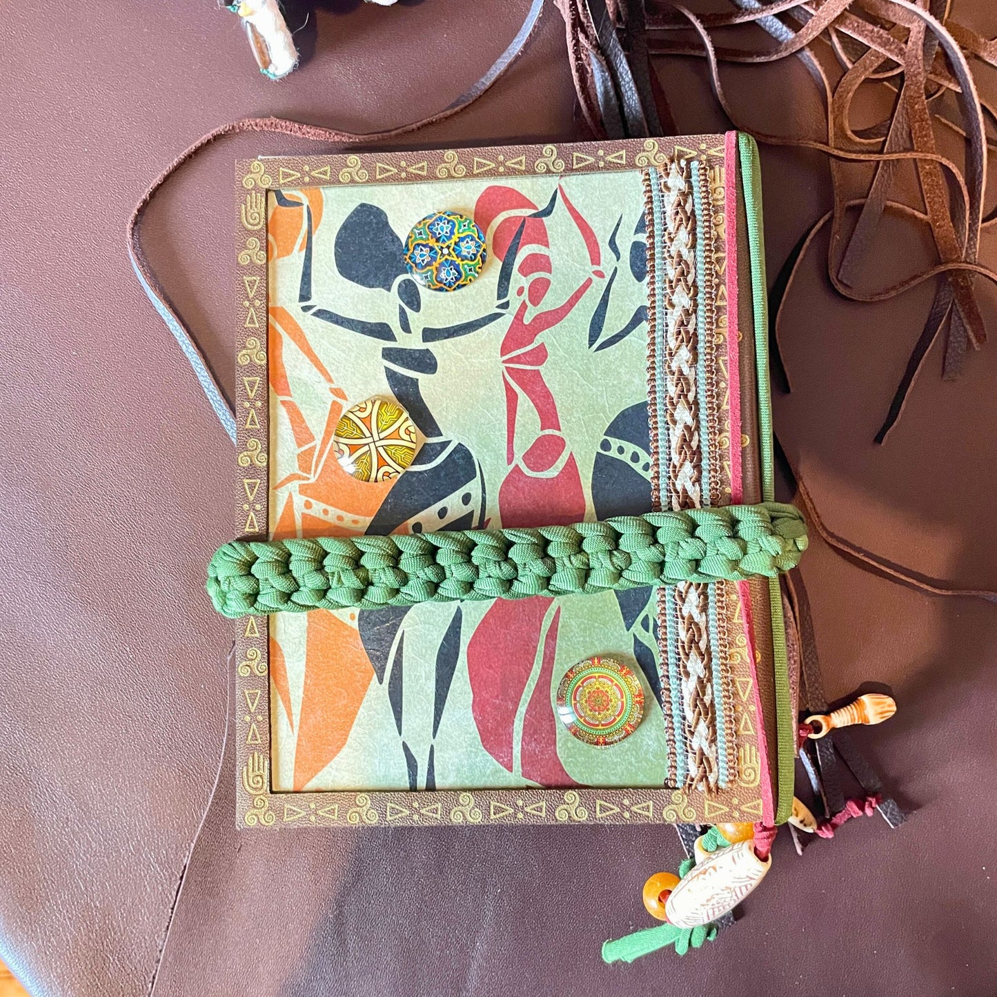 SteamVoyage - libro magico della sciamana in legno decorato con la tecnica del decoupage, rivestito in pelle marrone incisa al laser con fantasie africane dorate, con applicazioni in stoffa e perline e fettuccia per la chiusura in lycra lavorata all'uncinetto. All'interno, stampati su carta di pregio, si trovano preghiere e invocazioni, ricette per elisir magici e indicazioni per rituali arcani, oltre ad annotazioni personali della maga.