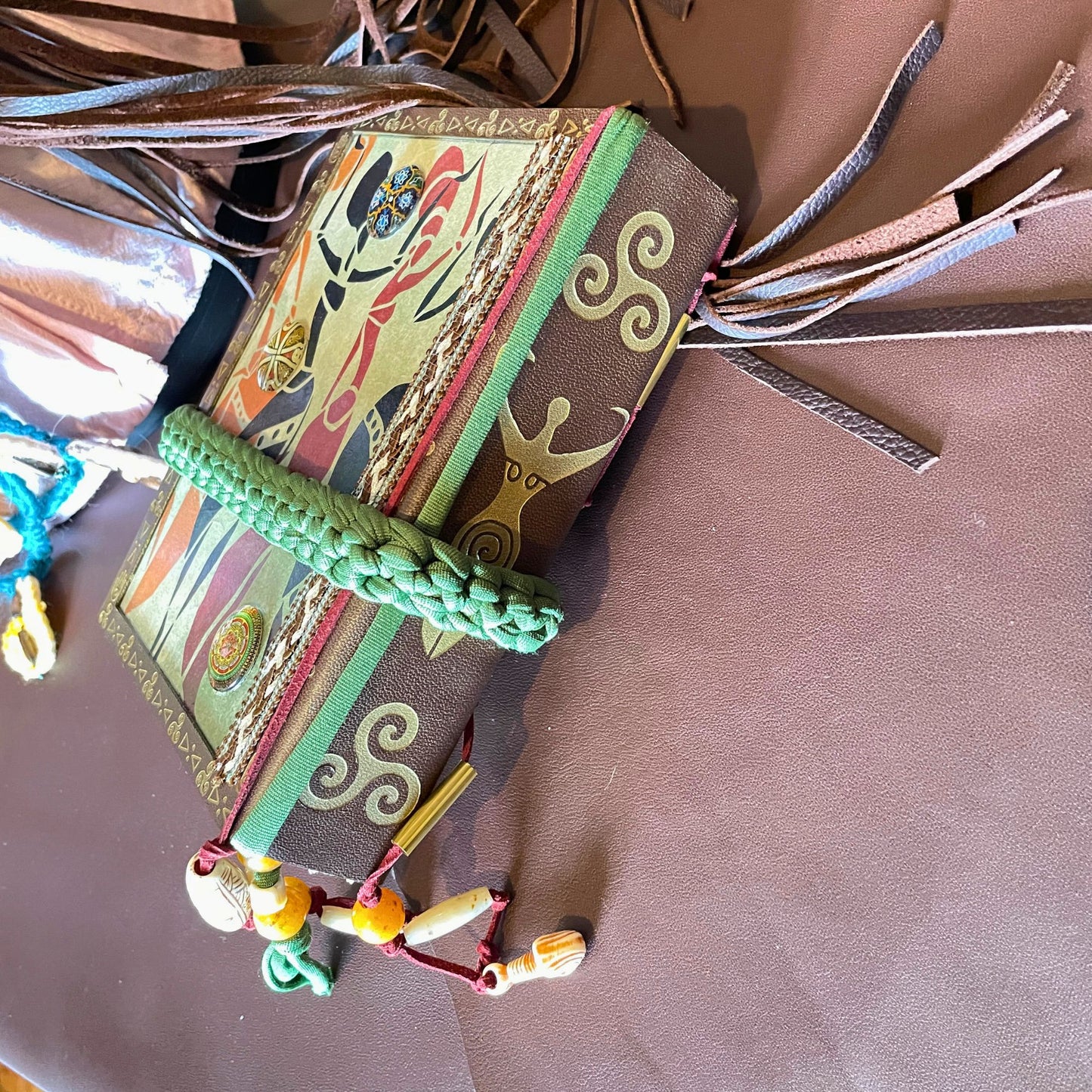 SteamVoyage - libro magico della sciamana in legno decorato con la tecnica del decoupage, rivestito in pelle marrone incisa al laser con fantasie africane dorate, con applicazioni in stoffa e perline e fettuccia per la chiusura in lycra lavorata all'uncinetto. All'interno, stampati su carta di pregio, si trovano preghiere e invocazioni, ricette per elisir magici e indicazioni per rituali arcani, oltre ad annotazioni personali della maga.