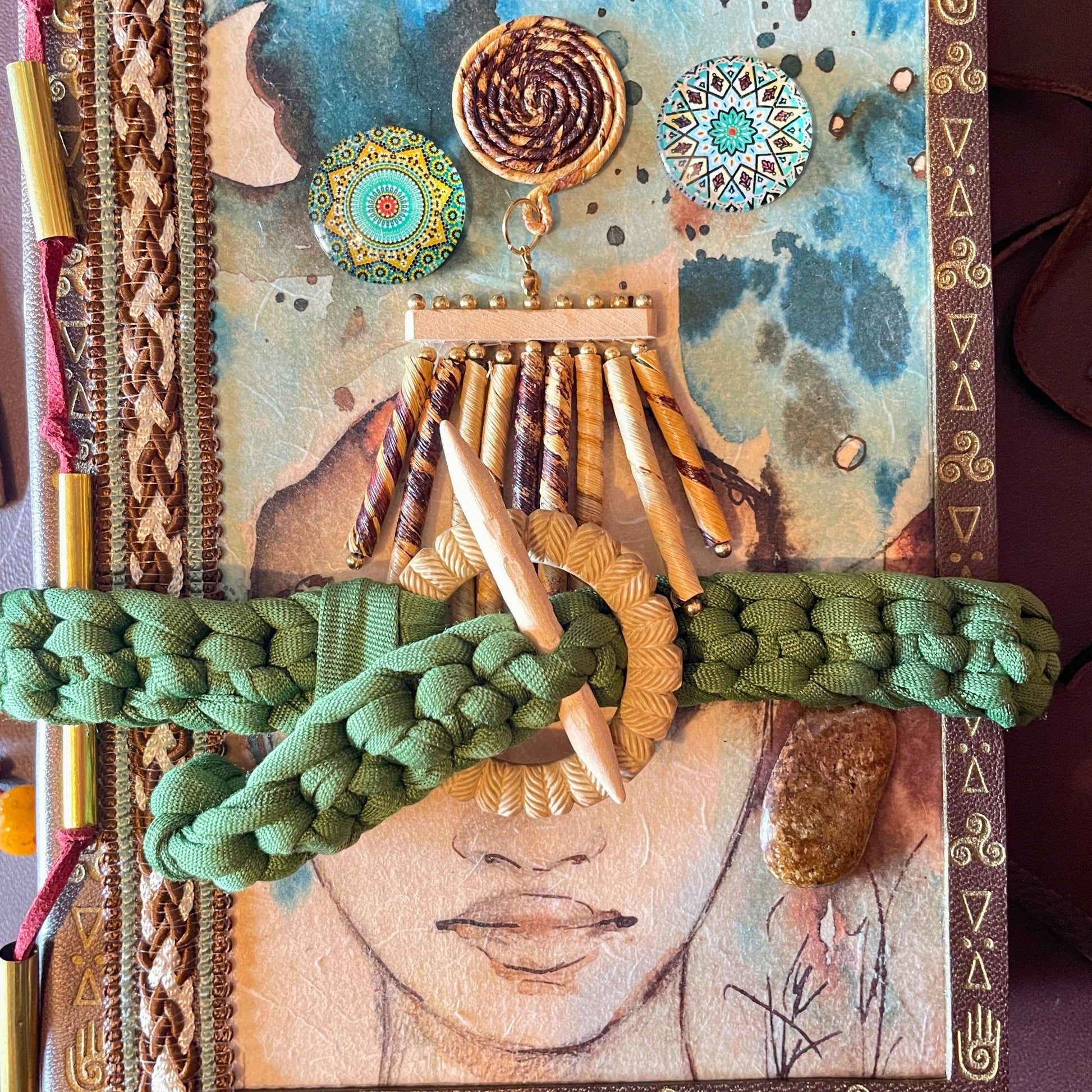 SteamVoyage - libro magico della sciamana in legno decorato con la tecnica del decoupage, rivestito in pelle marrone incisa al laser con fantasie africane dorate, con applicazioni in stoffa e perline e fettuccia per la chiusura in lycra lavorata all'uncinetto. All'interno, stampati su carta di pregio, si trovano preghiere e invocazioni, ricette per elisir magici e indicazioni per rituali arcani, oltre ad annotazioni personali della maga.