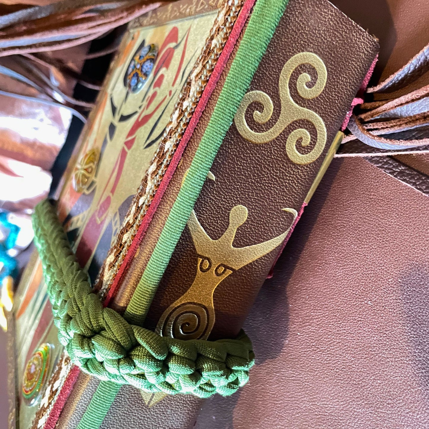 SteamVoyage - libro magico della sciamana in legno decorato con la tecnica del decoupage, rivestito in pelle marrone incisa al laser con fantasie africane dorate, con applicazioni in stoffa e perline e fettuccia per la chiusura in lycra lavorata all'uncinetto. All'interno, stampati su carta di pregio, si trovano preghiere e invocazioni, ricette per elisir magici e indicazioni per rituali arcani, oltre ad annotazioni personali della maga.