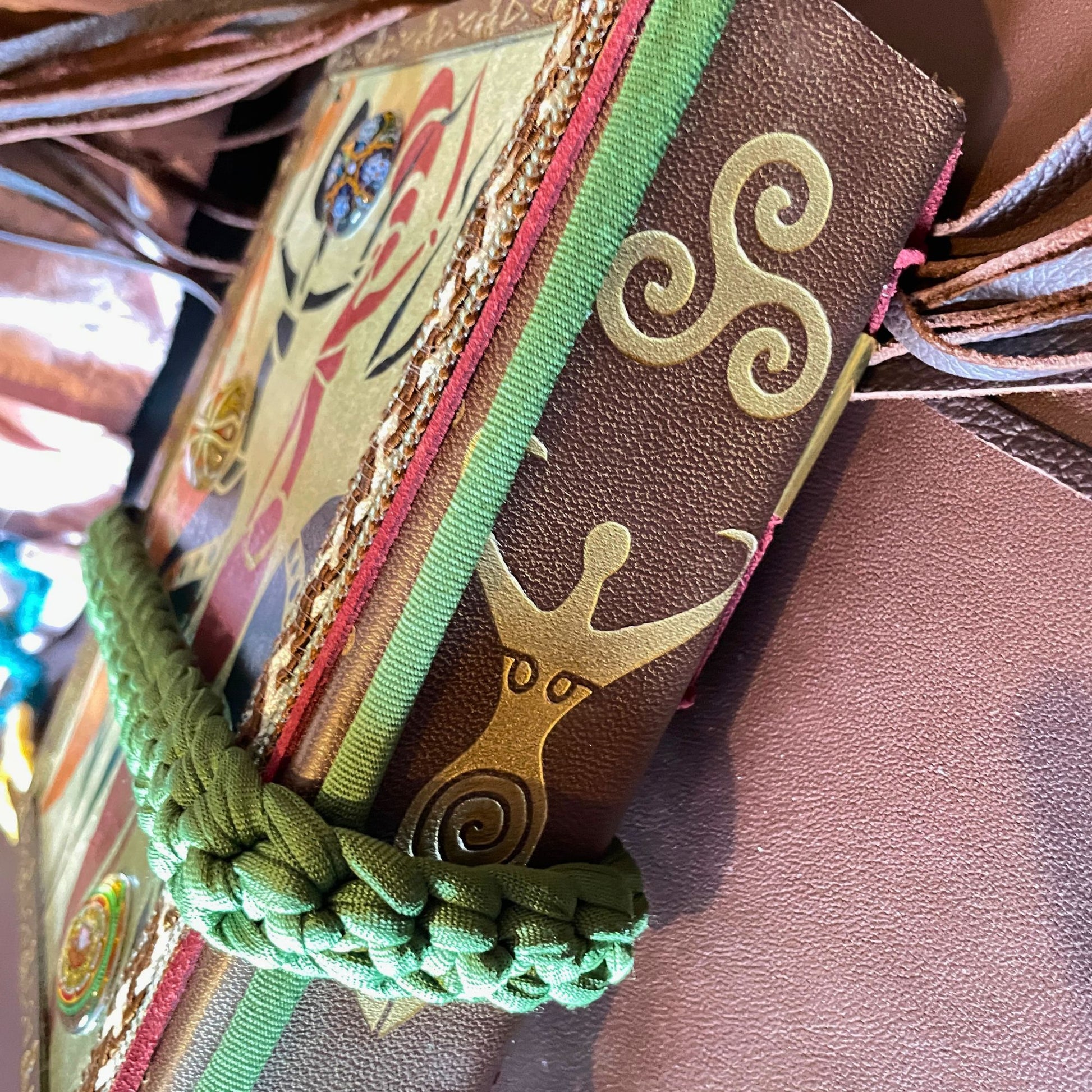 SteamVoyage - libro magico della sciamana in legno decorato con la tecnica del decoupage, rivestito in pelle marrone incisa al laser con fantasie africane dorate, con applicazioni in stoffa e perline e fettuccia per la chiusura in lycra lavorata all'uncinetto. All'interno, stampati su carta di pregio, si trovano preghiere e invocazioni, ricette per elisir magici e indicazioni per rituali arcani, oltre ad annotazioni personali della maga.