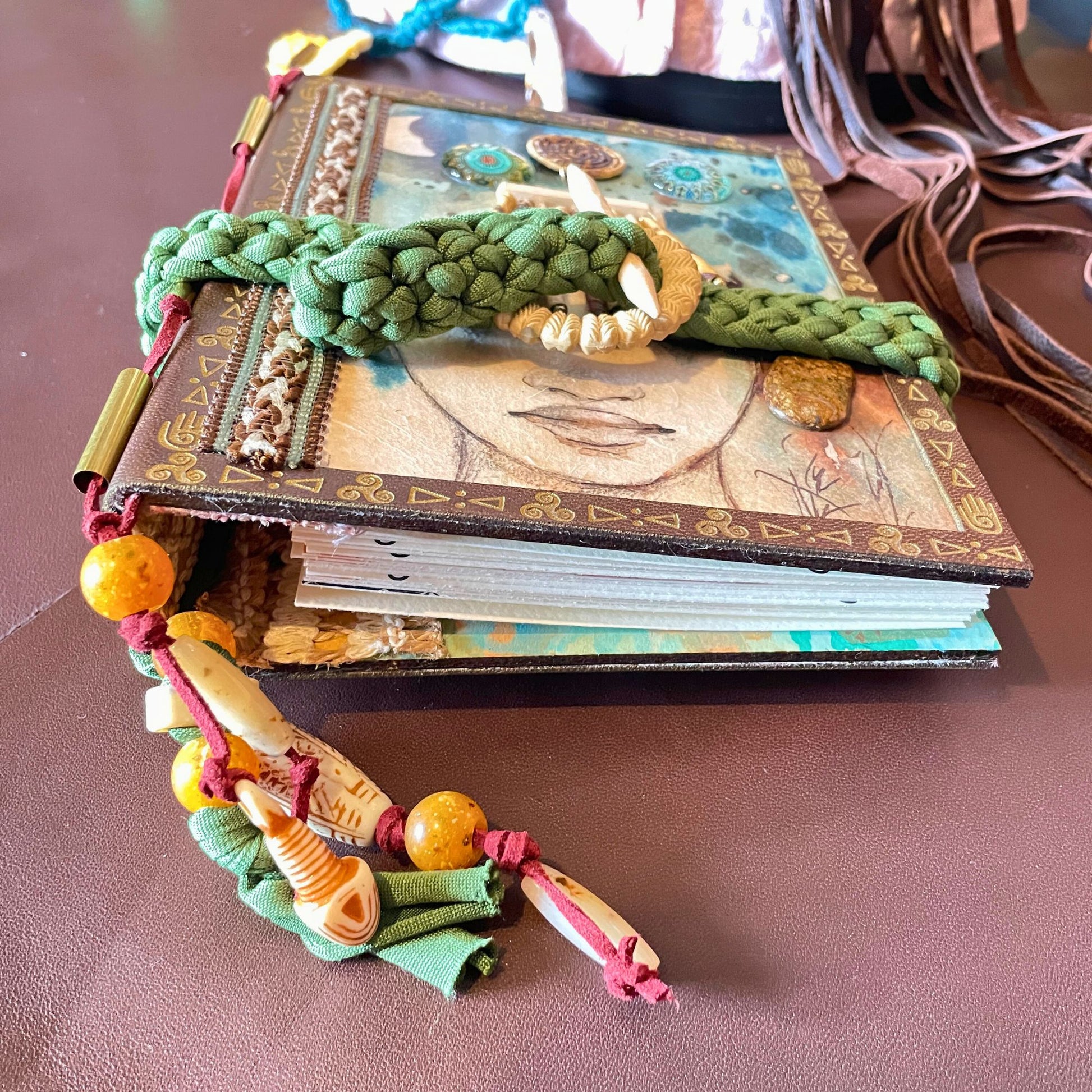 SteamVoyage - libro magico della sciamana in legno decorato con la tecnica del decoupage, rivestito in pelle marrone incisa al laser con fantasie africane dorate, con applicazioni in stoffa e perline e fettuccia per la chiusura in lycra lavorata all'uncinetto. All'interno, stampati su carta di pregio, si trovano preghiere e invocazioni, ricette per elisir magici e indicazioni per rituali arcani, oltre ad annotazioni personali della maga.