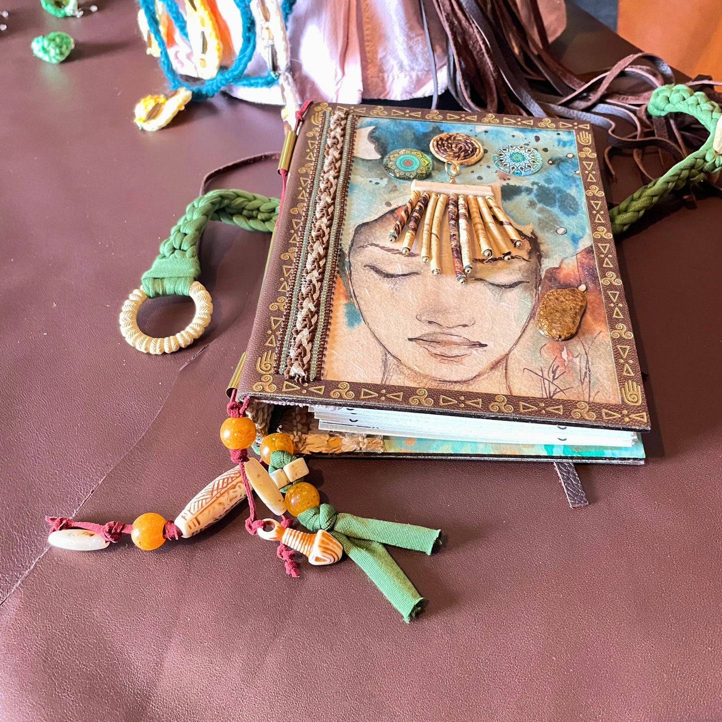 SteamVoyage - libro magico della sciamana in legno decorato con la tecnica del decoupage, rivestito in pelle marrone incisa al laser con fantasie africane dorate, con applicazioni in stoffa e perline e fettuccia per la chiusura in lycra lavorata all'uncinetto. All'interno, stampati su carta di pregio, si trovano preghiere e invocazioni, ricette per elisir magici e indicazioni per rituali arcani, oltre ad annotazioni personali della maga.