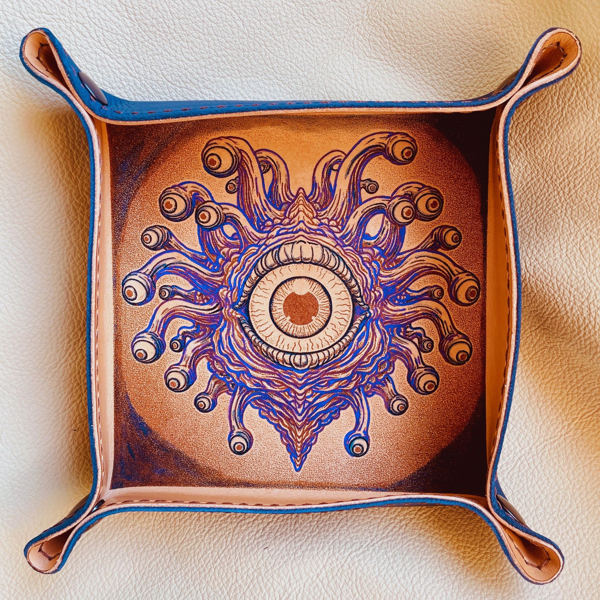 SteamVoyage - vassoio lancia dadi in cuoio dipinto a mano rivestito con pelle blu, dotato di quattro bottoni angolari per la chiusura e cucitura sul bordo. Il vassoio lancia dadi è decorato con l'immagine di un beholder, mostro tipico di Dungeons and dragons, dipinto con colori vivaci: viola e blu