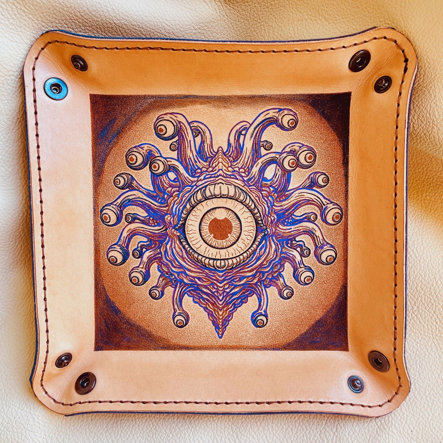 SteamVoyage - vassoio lancia dadi in cuoio dipinto a mano rivestito con pelle blu, dotato di quattro bottoni angolari per la chiusura e cucitura sul bordo. Il vassoio lancia dadi è decorato con l'immagine di un beholder, mostro tipico di Dungeons and dragons, dipinto con colori vivaci: viola e blu
