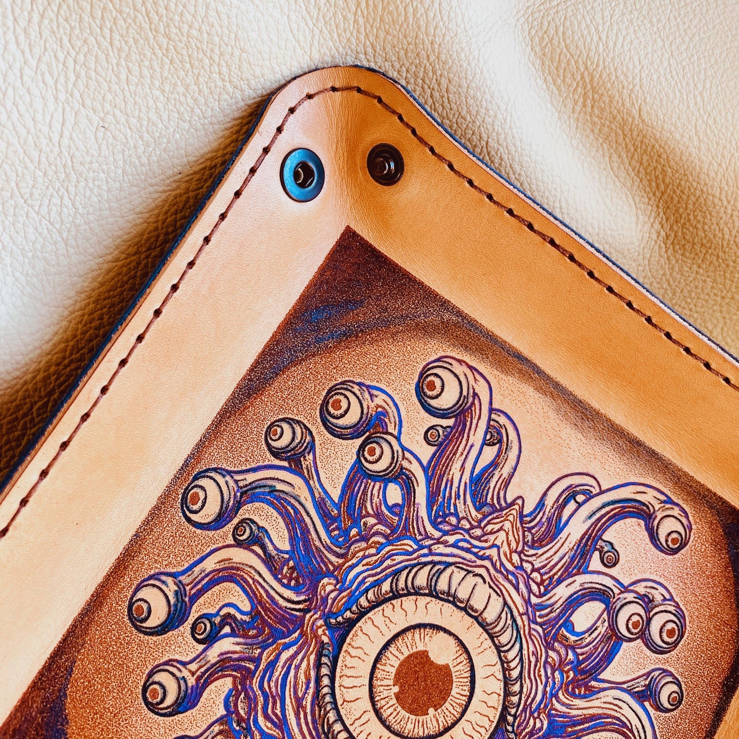 SteamVoyage - vassoio lancia dadi in cuoio dipinto a mano rivestito con pelle blu, dotato di quattro bottoni angolari per la chiusura e cucitura sul bordo. Il vassoio lancia dadi è decorato con l'immagine di un beholder, mostro tipico di Dungeons and dragons, dipinto con colori vivaci: viola e blu
